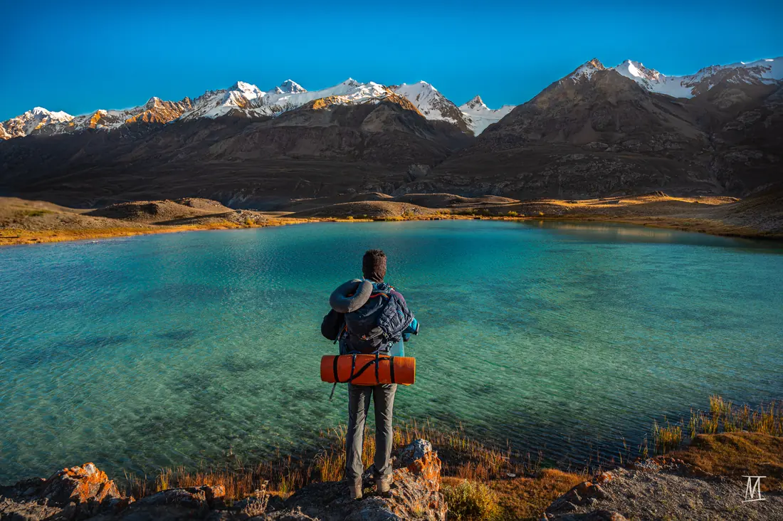 A trekker with a backpack stands at the edge of a turquoise alpine lake below snowy peaks